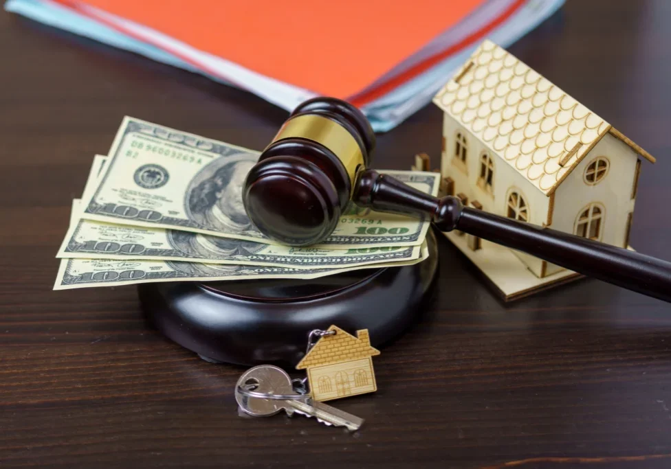 Gavel, money, house model, keys on table.